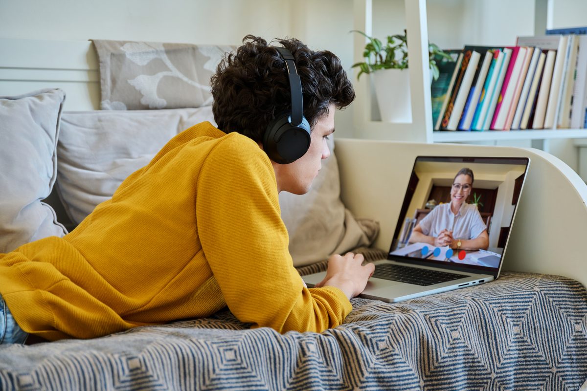 Junger Mensch mit Kopfhörern liegt auf dem Sofa und spricht per Video mit seiner Beraterin