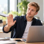 Junger Mann signalisiert am Arbeitsplatz mit erhobener Hand ein Nein, während Kollegen ihm weitere Aufgaben anbieten. Die Szene zeigt, wie wichtig es ist, persönliche Grenzen zu setzen.